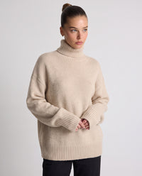 A woman wearing a beige ribbed turtleneck sweater with oversized sleeves, paired with black pants, standing against a light gray studio background.